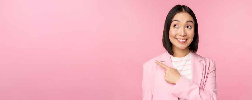 Enthusiastic young asian businesswoman, office employee pointing finger left, looking at banner, advertisement with happy smile, showing advertisement, pink background.