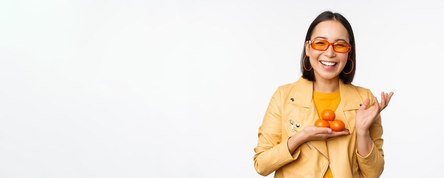 Stylish happy asian girl in sunglasses holding tangerines and smiling, posing against white background. Copy space