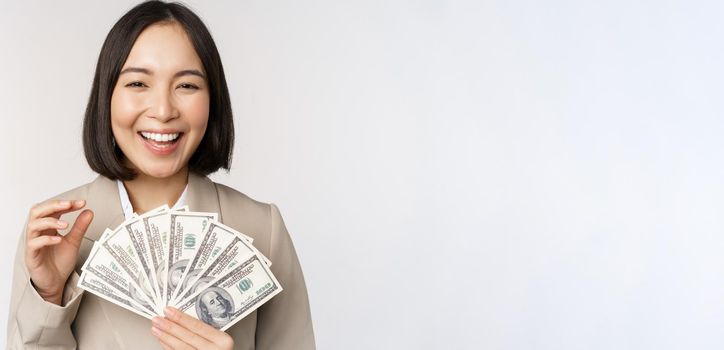 Image of successful businesswoman holding money. Asian corporate woman with cash dollars, smiling and laughing, standing over white background.