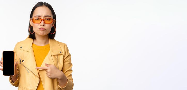 Portrait of sad asian woman in sunglasses, pointing finger at mobile phone app interface, showing smartphone application, standing over white background.