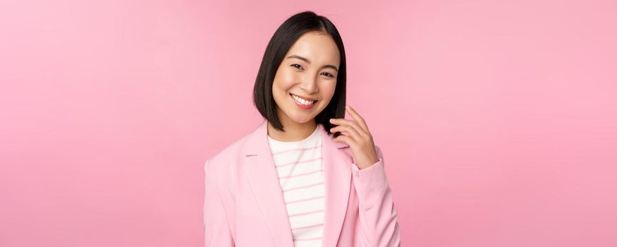 Young professionals. Smiling asian businesswoman, saleswoman in suit looking confident at camera, posing against pink background.