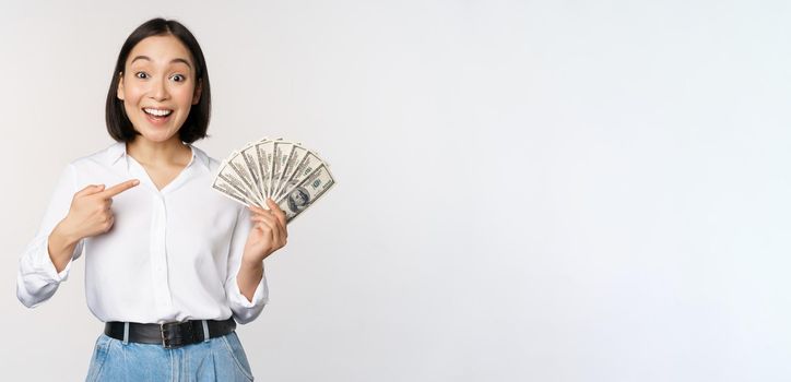 Portrait of young asian woman pointing at her money dollars, showing cash, standing over white background.