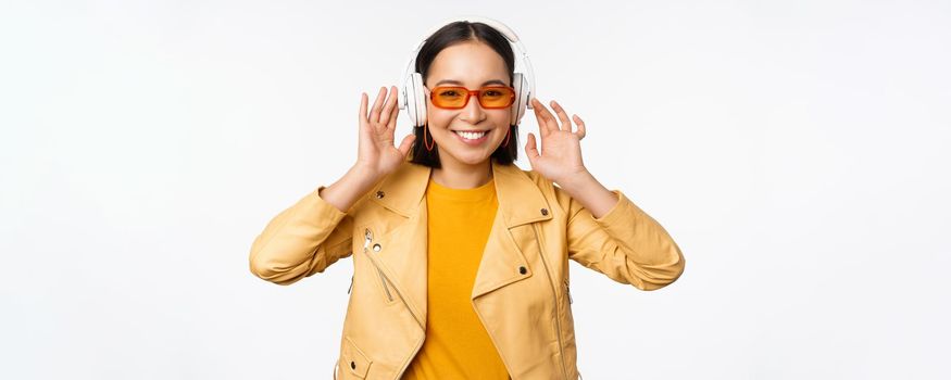 Beautiful asian girl, laughing happy, listening music in headphones, standing over white background. Copy space