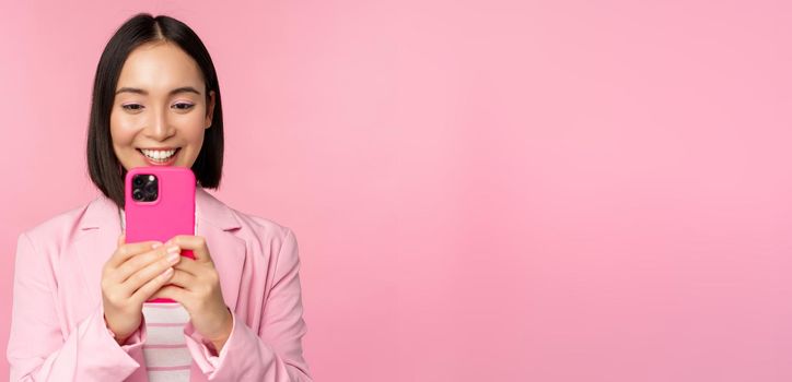 Image of smiling asian corporate woman in suit looking, watching on smartphone app, using mobile phone application, standing over pink background.