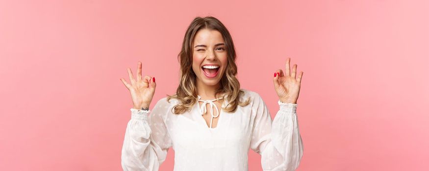 Close-up portrait of beautiful blond girl in white dress guarantee you will enjoy this event, show okay sign, confirm everything excellent, wink and smiling carefree, standing pink background.