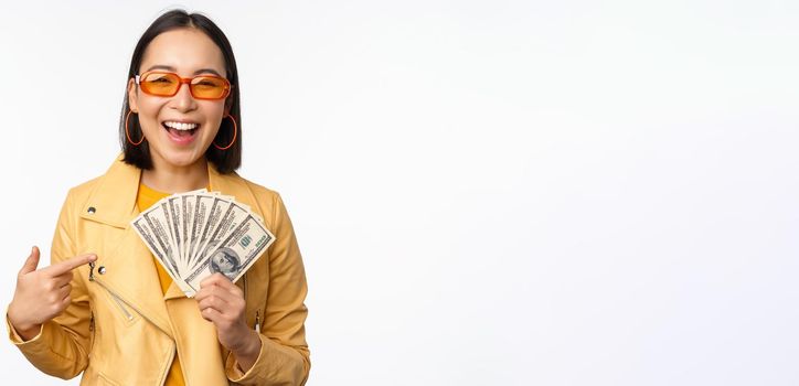 Microcredit and money concept. Stylish asian young woman in sunglasses, laughing happy, holding dollars cash, standing over white background.