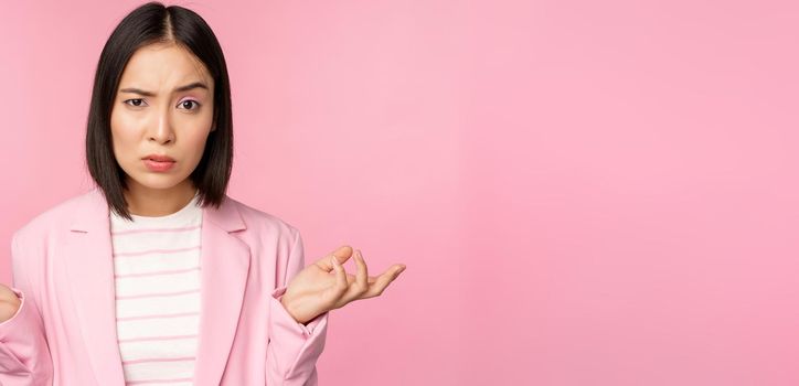 Portrait of young asian business woman, saleswoman shrugging shoulders and looking confused, clueless of smth, standing over pink background.