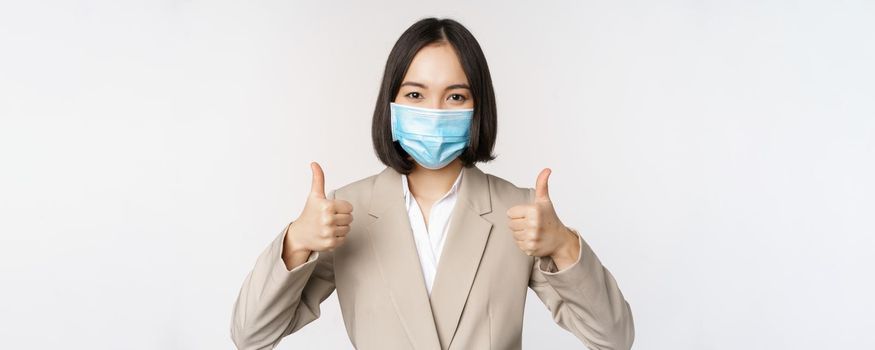 Coronavirus and workplace concept. Image of asian saleswoman, company worker in medical mask showing thumbs up sign, smiling pleased, white background.