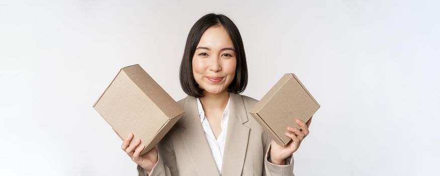 Image of saleswoman, asian businesswoman holding boxes with company brand product, smiling at camera, standing against white background.