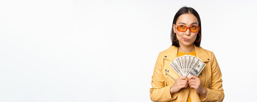 Microcredit and money concept. Stylish asian young woman in sunglasses, laughing happy, holding dollars cash, standing over white background.