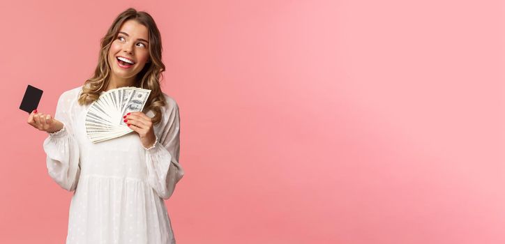 Portrait of pleased attractive blond girl in white dress, daydreaming about what buy, shopping with girlfriends, holding money and credit card, use cash, standing pink background.