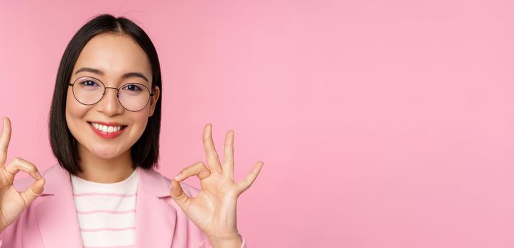 Close up portrait of impressed corporate woman, asian business lady in glasses, showing okay sign, looking amazed at camera, recommending, pink background.