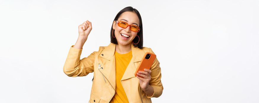 Enthusiastic korean girl in sunglasses, holding smartphone, celebrating and dancing, laughing happy with mobile phone, standing over white background.