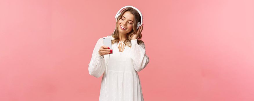 Portrait of dreamy cute and tender blond girl in white dress, holding smartphone, listen music in headphones, smiling at display mobile phone as picking playlist for spring mood, pink background.