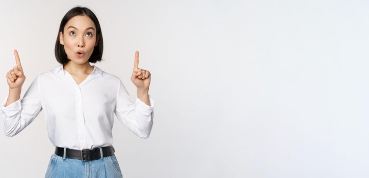 Enthusiastic asian business woman pointing, looking up with happy smiling face, showing company logo or banner, standing over white background.