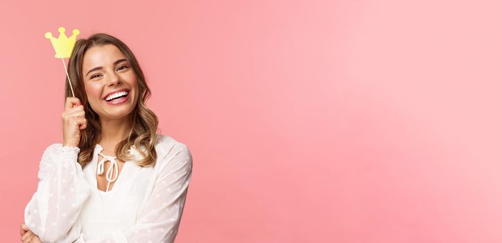 Spring, happiness and celebration concept. Close-up portrait of charming smiling, lovely blond girl holding small queen crown on stick, laughing joyfully, feel empowered and happy, pink background.