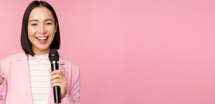 Image of enthusiastic asian businesswoman giving speech, talking with microphone, holding mic, standing in suit against pink studio background.