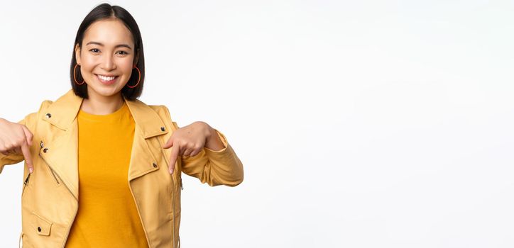 Portrait of happy smiling asian girl, pointing fingers down and showing logo, demonstrating banner, standing in yellow jacket against white background. Copy space