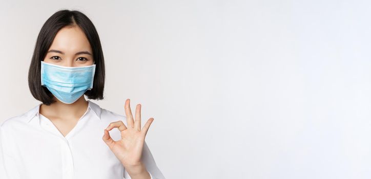 Covid and health concept. Portrait of asian woman wearing medical face mask and showing okay sign, standing over white background.