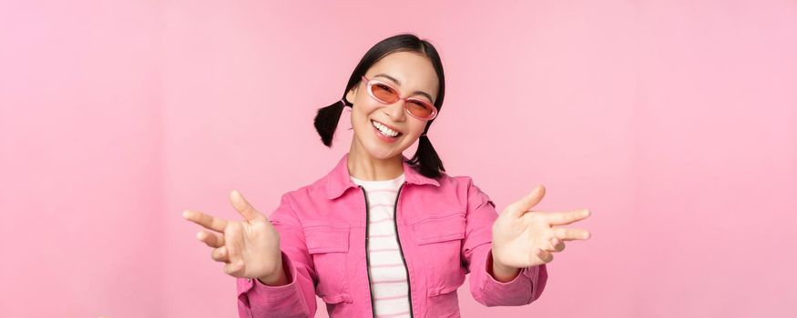 Happy stylish korean girl in sunglasses, reaching hands, receiving, smiling friendly and welcome you, standing over pink background.