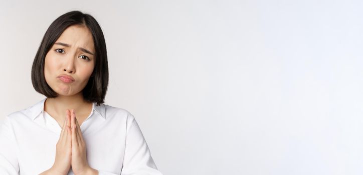 Close up portrait of asian woman begging, asking for smth, say please with coy sad face, need help, standing over white background.