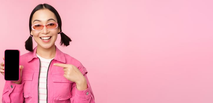 Enthusiastic asian woman in stylish clothes, sunglasses, pointing finger at mobile phone screen, showing smartphone application, standing over pink background.