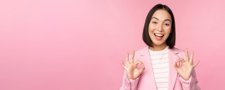 Okay, excellent. Businesswoman in corporate suit, showing ok, approval gesture, recommending smth, give positive feedback and smiling pleased, posing over pink background.