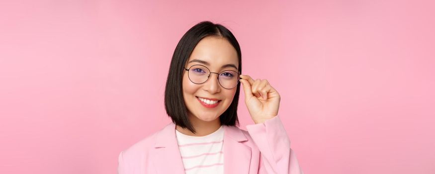 Successful asian businesswoman in glasses, smiling and looking professional at camera, wearing suit, standing over pink background.