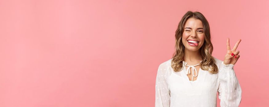 Close-up portrait of friendly joyful blond girl in white dress, sending positive vibes, smiling pleased and wink, stick tongue showing peace sign, looking kawaii over pink background.