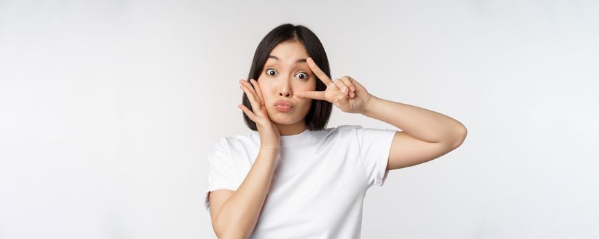 Cute asian girl posing with kawaii v-sign, peace gesture near face, standing in tshirt over white background. Copy space