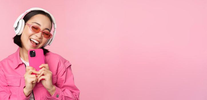 Stylish asian girl in headphones, listening music and taking photos on mobile phone, using smartphone, standing over pink background.