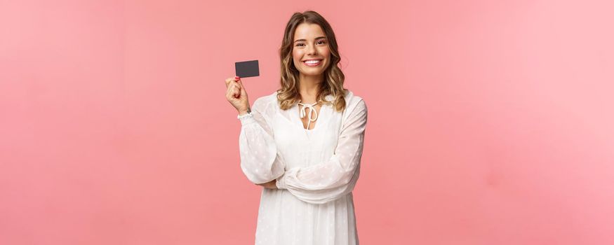 Portrait of pleased good-looking blond european female in white dress, show credit card with satisfied expression, smiling camera, recommend bank services, use payment online, pink background.