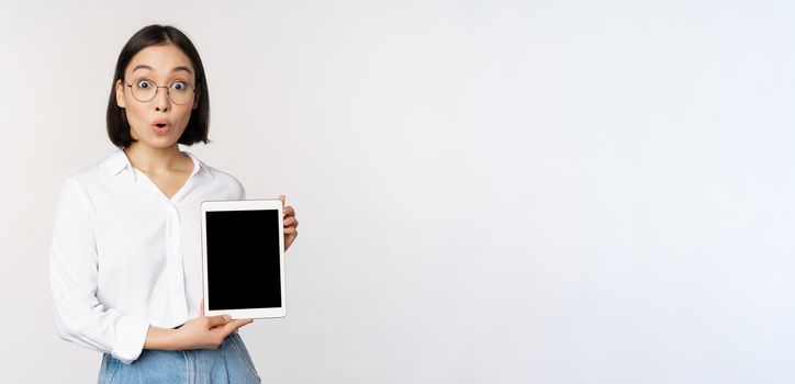 Enthusiastic asian woman, office worker in glasses showing digital tablet screen, demonstrating info on gadget display, standing over white background.