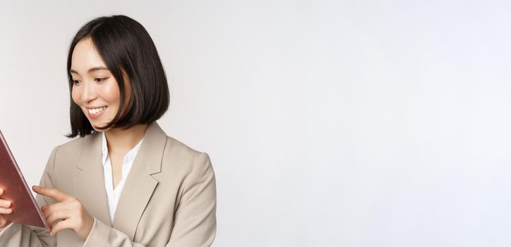 Image of asian businesswoman using digital tablet, looking at gadget and smiling, working, standing against white background.