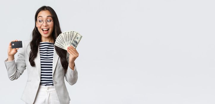 Business, finance and employment, entrepreneur and money concept. Indecisive cute asian office lady thinking about putting cash on deposit, looking excited at credit card, white background.