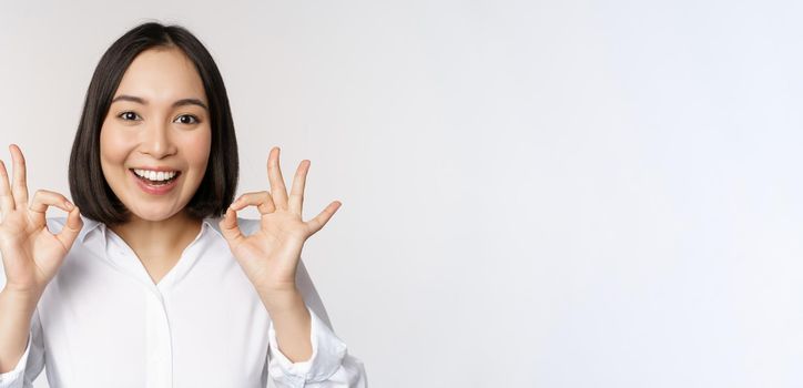 Close up head portrait of asian girl showing okay, ok sign and smiling satisfied, recommending, being pleased, praise and make compliment, white background.