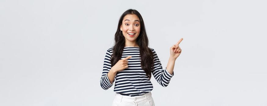 Lifestyle, beauty and fashion, people emotions concept. Surprised happy asian girl standing over white background recommend visit shop, pointing fingers right at banner.