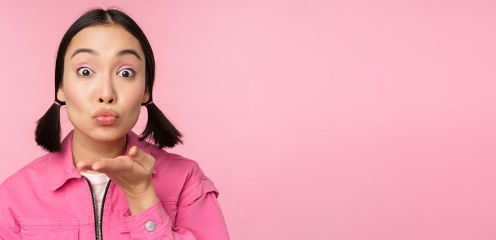 Close up portrait of beautiful silly asian girl, sending air kiss, mwah at camera, blowing, standing over pink background.