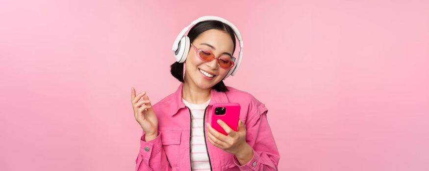 Stylish asian girl dancing with smartphone, listening music in headphones on mobile phone app, smiling and laughing, posing against pink background.