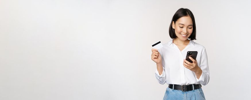 Online shopping concept. Korean woman holding credit card and looking at smartphone app, buying, order delivery in mobile phone application, standing over white background.