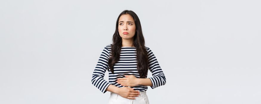 Lifestyle, beauty and fashion, people emotions concept. Woman got food poisoned, touching belly feeling unwell. Asian girl with cramps looking gloomy, having menstrual pain, white background.