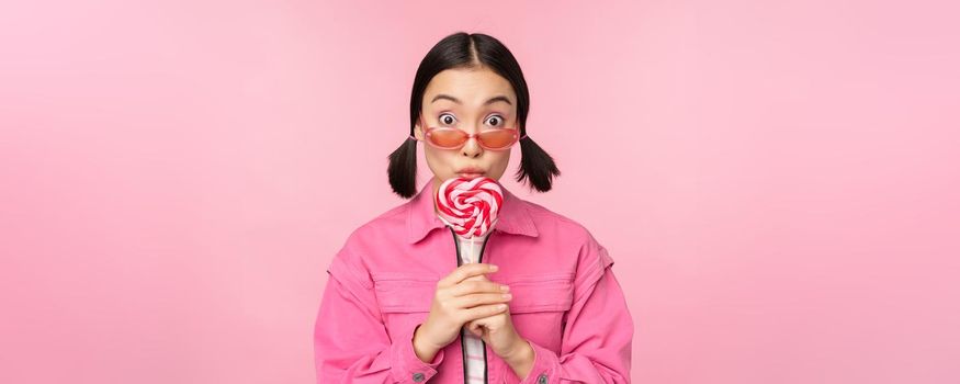 Silly and cute asian female model licking lolipop, eating candy sweet and smiling, looking excited, standing over pink background.