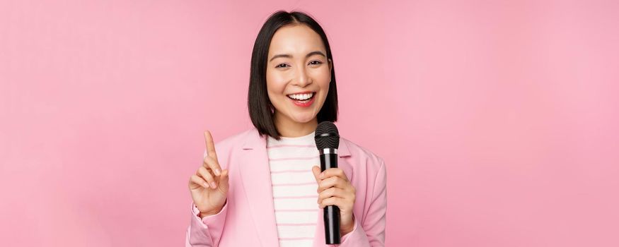 Image of enthusiastic asian businesswoman giving speech, talking with microphone, holding mic, standing in suit against pink studio background.