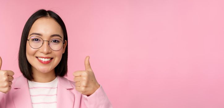 Awesome, congrats. Face of excited asian businesswoman in glasses, smiling pleased, showing thumbs up in approval, standing over pink background.