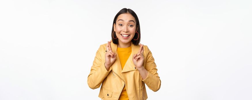 Portrait of excited asian woman looks hopeful, wishing, praying or begging, waiting for news, standing over white background, smiling enthusiastic.