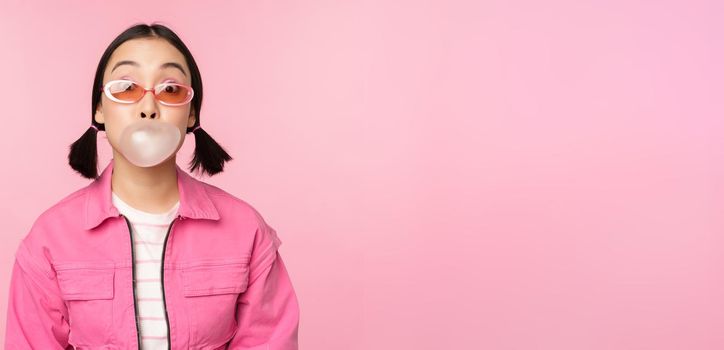Stylish asian girl blowing bubblegum bubble, chewing gum, wearing sunglasses, posing against pink background. Copy space