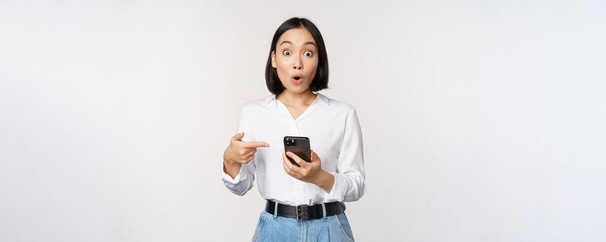 Amazed girl talking about smartphone app, pointing at phone while looking impressed at camera, standing against white background.