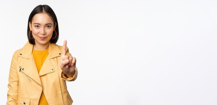 Rule number one. Confident smiling asian woman showing index finger, stop, prohibit gesture, disapprove smth, standing over white background.