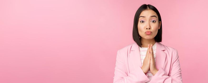 Portrait of asian businesswoman asking for help, say please, standing in praying, begging pose, pink studio background.