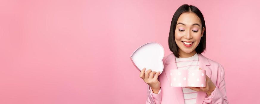 Happy cute korean girl in suit, opens up heart shaped box with romantic gift on white day holiday, standing in suit over pink background.